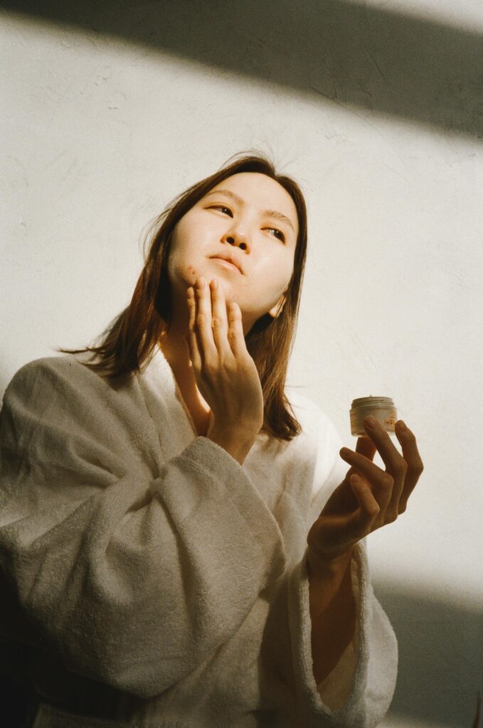 Asian woman in bathrobe applying moisturizer for her skincare routine. Bright sunlight illuminates the scene.